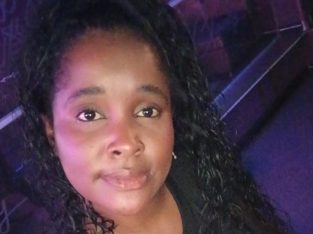 Fer69s has dark curly hair, brown eyes, and a slight smile with shiny lips, wearing a black shirt, looking straight at the camera with a relaxed expression.