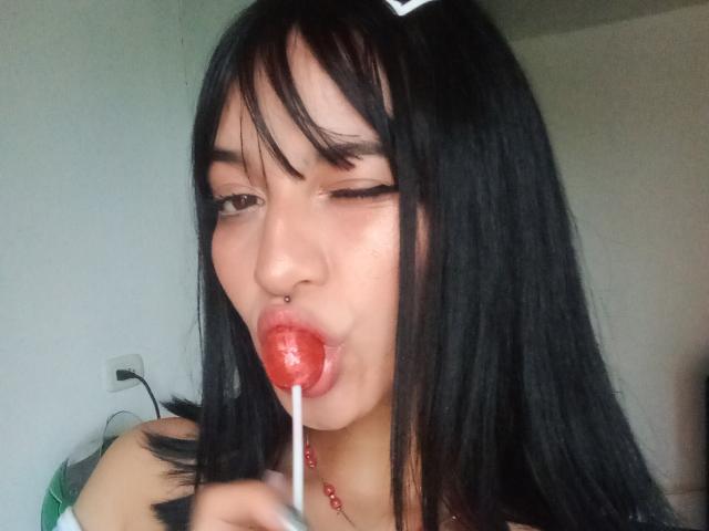LindaWatsong with black hair, winking, sucking a red lollipop, nose pierced, holding the stick in her right hand, wearing a red bead necklace.