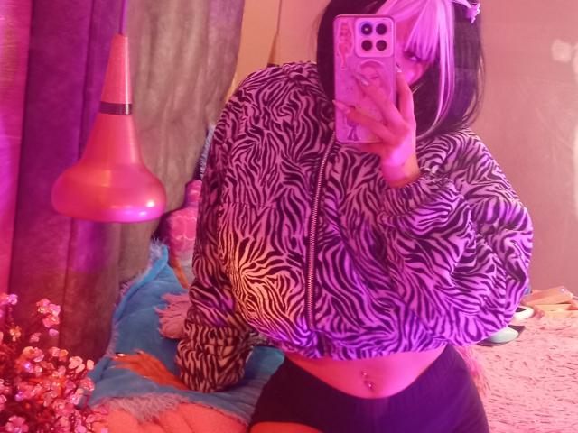 LindaWatsong with huge tits, zebra print hoodie, black and white hair, mirror selfie, pink phone, belly button piercing, black shorts, pink lamp, blue pillow, and flowers.