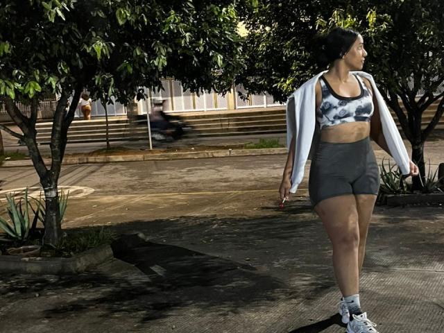 ThiainaRose, met donkere huid, draagt een zwarte en witte sportbeha, grijze shorts, witte sokken en witte sneakers. Ze loopt, met een witte jas die over haar schouders hangt..