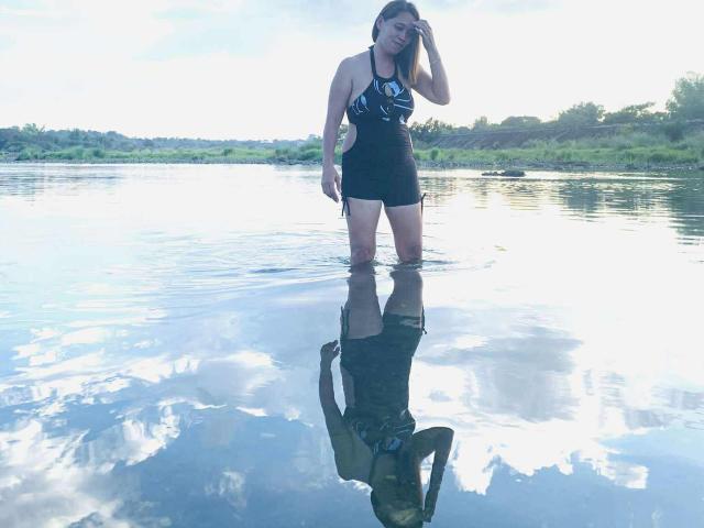 Kath69 stands in shallow water, wearing a black and blue one-piece swimsuit, her reflection visible in the clear water, with her hand in her brown hair.