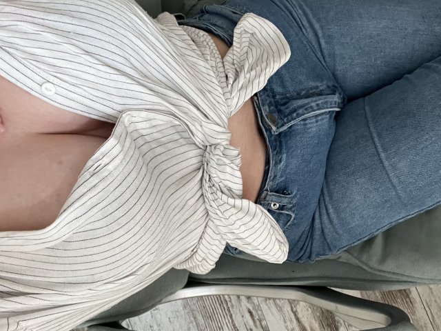 Rimia's tits are showing through her white, striped, button-up shirt tied at the front. She's wearing tight blue jeans, sitting on a green chair.