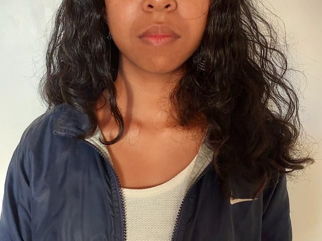 Rosiia, met een medium bruine huid, golvend zwart haar, draagt een marineblauwe jas over een witte Nike shirt, waarbij haar bovenborst en gezicht zichtbaar zijn, met licht open lippen..