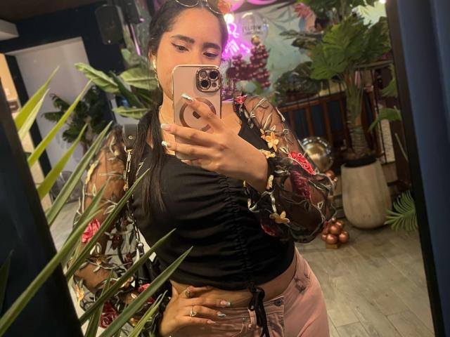SiennaTaylor with long black hair, wearing a black floral top, pink pants, holding a phone in a mirror selfie, showing her tits and hand with nails.
