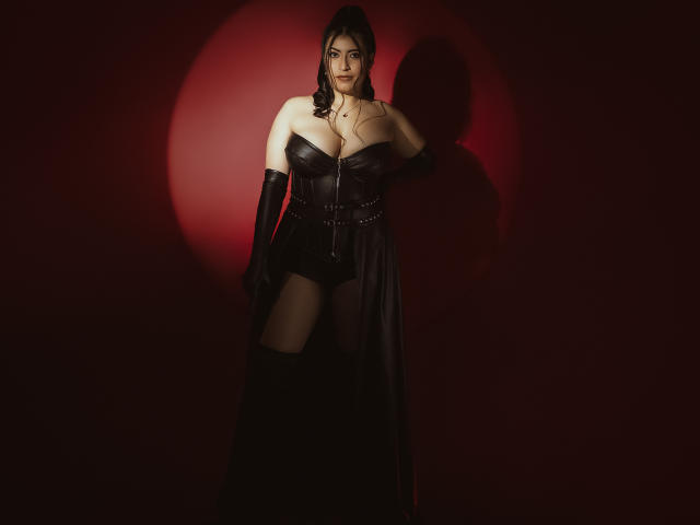 KamileNilsen with big tits, wearing a black leather corset, gloves, and thigh-high boots, standing against a red circle, looking seductive, dark hair, pale skin, serious expression.