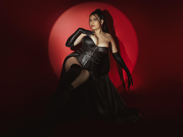 KamileNilsen with black leather corset, gloves, and dress, showing her big boobs and thick thighs, sitting against a red circle, looking seductive, long black hair in a ponytail.