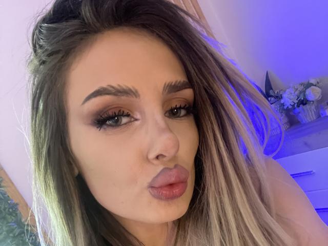 RheaI has pouty lips, thick eyebrows, long eyelashes, and blonde-to-brown ombre hair in a selfie. Her tits are partially visible. She's making a pouty face.
