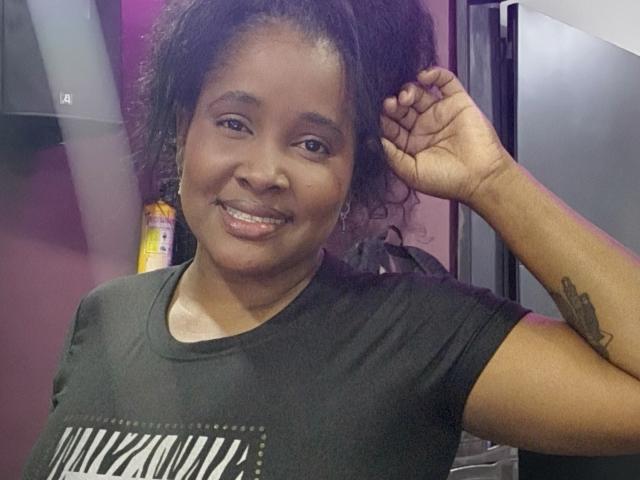 Fer69s with dark skin, curly black hair, small nose, smiling with slightly parted lips, wearing a black zebra-print shirt, right hand in hair, visible arm tattoo.