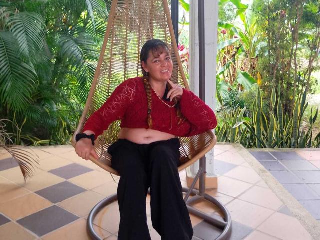 AmandaHotL with braided hair, red shirt, black pants, sitting in a wicker chair, hand on chin, slightly chubby belly, black bracelet, smiling, green foliage in background.