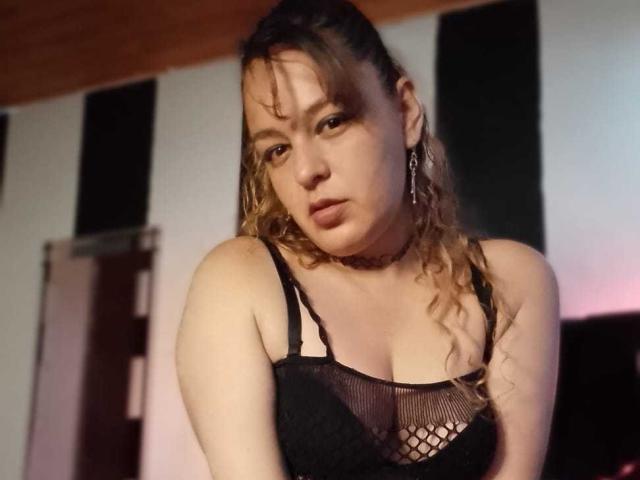 AmandaHotL with curly blonde hair, black mesh bra, showing her tits, choker, and earrings, looking slightly annoyed, in a dimly lit room.