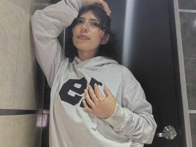 ToryPussCaty met donker krullend haar, die een grijze hoodie draagt met een zwart "P"-logo, met een hand in haar haar en de andere hand op haar borst, staand voor een zwarte deur..