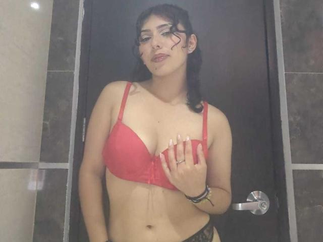 ToryPussCaty, avec des cheveux foncés, un soutien-gorge rouge et des culottes en dentelle noire, une main sur la poitrine, portant des bagues et des bracelets, debout devant une porte sombre. De l'eau coule du soutien-gorge..