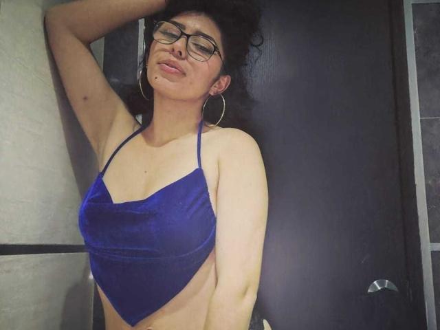 ToryPussCaty met krullend donker haar, een bril en oorringen, draagt een blauwe fluwelen haltertop, waardoor haar borsten en zijkant zichtbaar zijn, haar arm is omhoog, haar lippen zijn lichtjes open en haar ogen zijn gesloten..