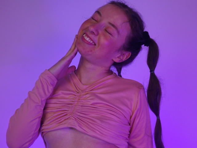 VioletaNova with a happy smile, eyes closed, pink ruched top, long brown pigtails, hand on cheek, purple-lit background, visible freckles, medium breasts.