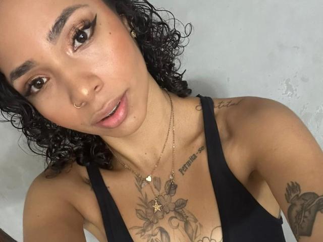 SilvanaXOwen has curly black hair, a nose ring, and wears a black tank top. She has tattoos on her chest and arm, and a gold necklace with a star.
