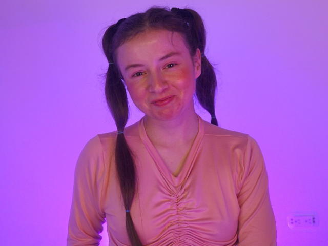 VioletaNova has pigtails, a cute smile, and is wearing a pink, ruched long-sleeve top. She has a slight red mark on her right cheek. Purple lighting.