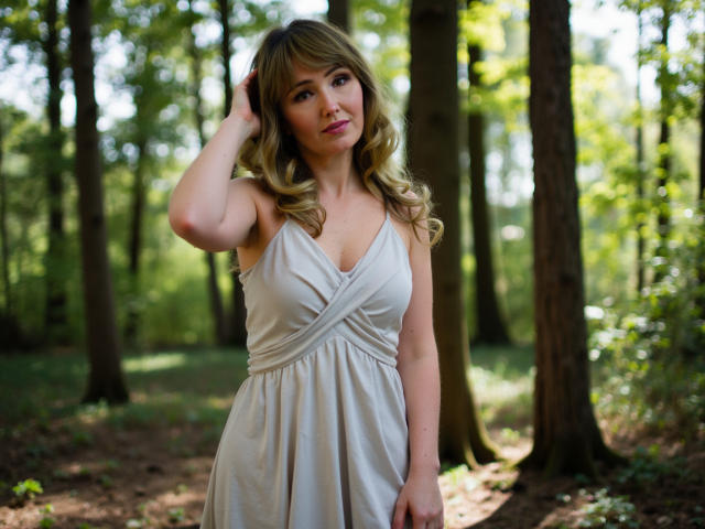 Justine, met golvend blond haar, draagt een witte jurk en staat in een bos. Ze raakt haar hoofd aan met een lichte glimlach, terwijl ze haar borsten laat zien en een schattig gezicht heeft..
