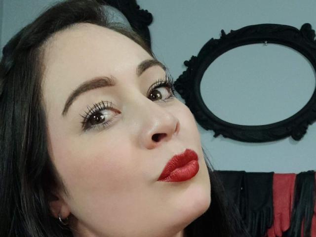 Il viso di Kristel Rouse, pelle chiara, sopracciglia scure, ciglia lunghe, rossetto rosso, capelli neri, testa leggermente inclinata, sguardo rivolto verso l'alto, abito nero e rosso sullo sfondo..