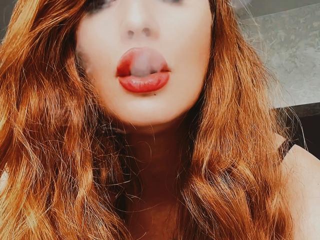 MayaGloss hat rote Lippen, die leicht geöffnet sind und ihre Zunge zeigen. Sie hat welliges, rotes Haar, das ihre Schultern bedeckt. Ihre helle Haut ist sichtbar, und sie schaut nach unten in die Kamera..