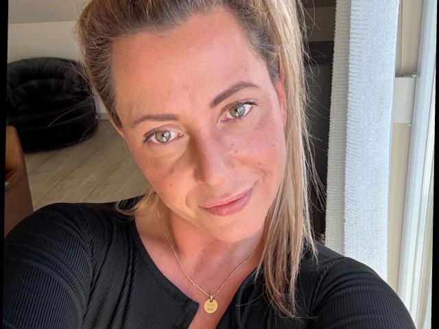 DeutscheLadyLena - aantrekkelijk met groene ogen, een lichtbruine paardenstaart, een lichte glimlach, gekleed in een zwart geribbeld shirt, een gouden ketting, een door de zon verlicht gezicht, zachte huid, subtiele sproeten en schattige roze lippen..
