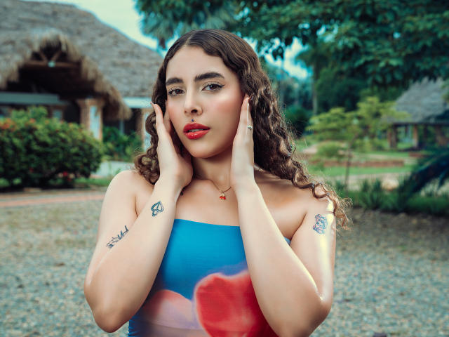 TianaVera with curly brown hair, red lipstick, blue strapless top, hands on cheeks, tattoos on forearms, standing outside, gravel ground, wooden hut in background, green trees.