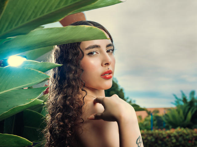 TianaVera with curly brown hair, red lipstick, and a tattoo on her upper arm, holding her chin, looking over her shoulder, surrounded by green leaves and a blue light.