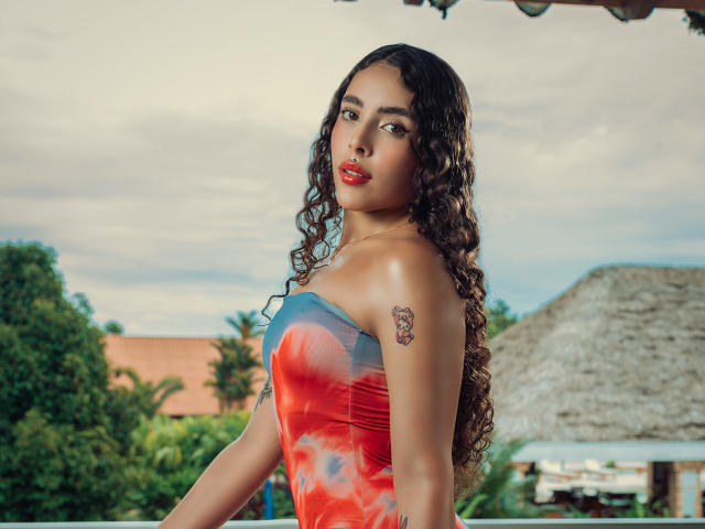 TianaVera with curly brown hair, red lipstick, and a colorful strapless top, showing a tattoo on her right shoulder, standing outside with a relaxed, confident expression.