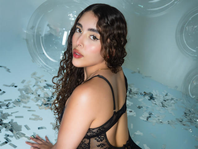 TianaVera with curly brown hair, looking over her shoulder, wearing black lace lingerie, showing her back and butt, sitting on a floor with scattered silver pieces.