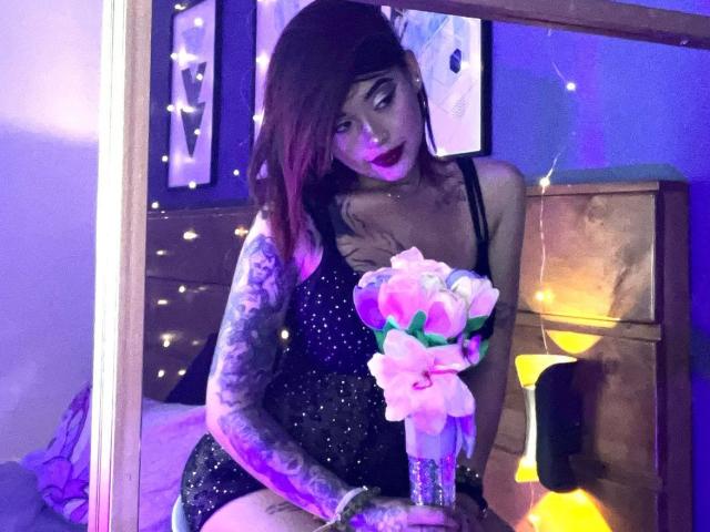 LolitaRose with tattoos, dark lipstick, and red hair, wearing a sparkly black dress, holding pink roses, sitting in a bedroom with purple and yellow lights.