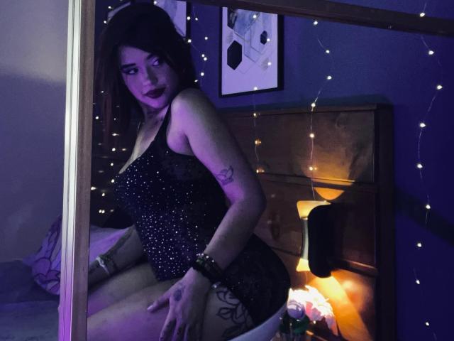 LolitaRose with dark makeup and purple highlights, wearing a sparkly black bodysuit, sitting on a bed, showing off her tattooed thigh and black bracelet, surrounded by fairy lights.
