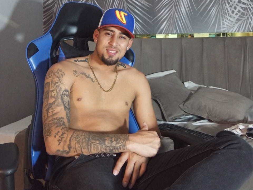 RockyMasterr, shirtless with tattoos, wears a blue and yellow cap, black pants, and a gold chain, sitting in a blue gaming chair, smiling.