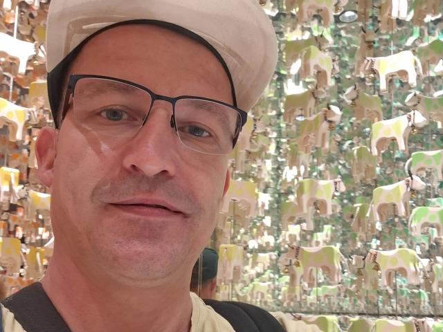 Tom Stern, met bril, witte hoed en een lichte snor, glimlacht naar de camera. Hij heeft een glanzende neus en draagt een zwart en beige shirt..