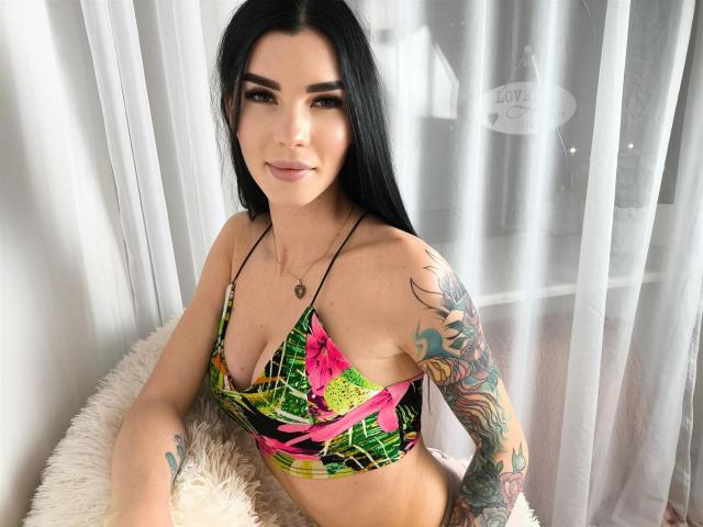 SofiaDream with long black hair, wearing a colorful floral bikini top, showing her tattooed arm and small tits, sitting on a fluffy white chair, smiling softly.