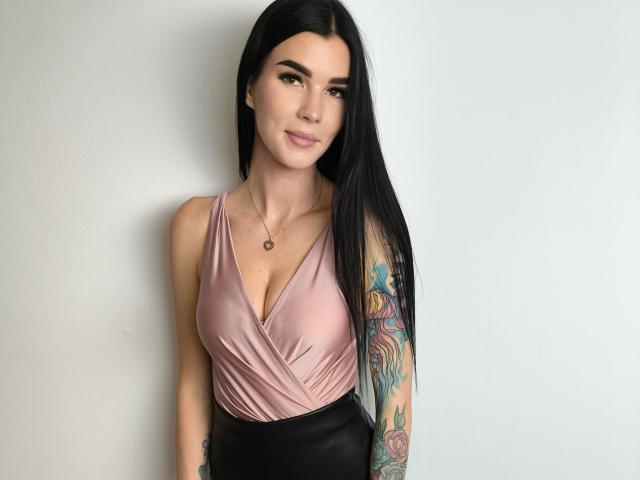 SofiaDream with long black hair, pink satin top, black leather skirt, tattooed arm, small necklace, smiling, showing her tits, standing against a white wall.