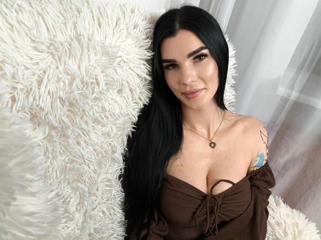 SofiaDream with long black hair, brown off-shoulder top, heart necklace, tattoo on right shoulder, sitting on fluffy white couch, smiling at camera, showing her tits.
