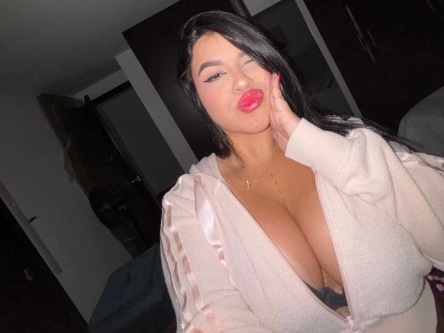 mujer atractiva, con senos grandes, cabello negro, labios rojos, vestida con una bata de satén rosa, guiñando un ojo, con la mano en la mejilla, en una habitación con poca luz, luz suave en su rostro..