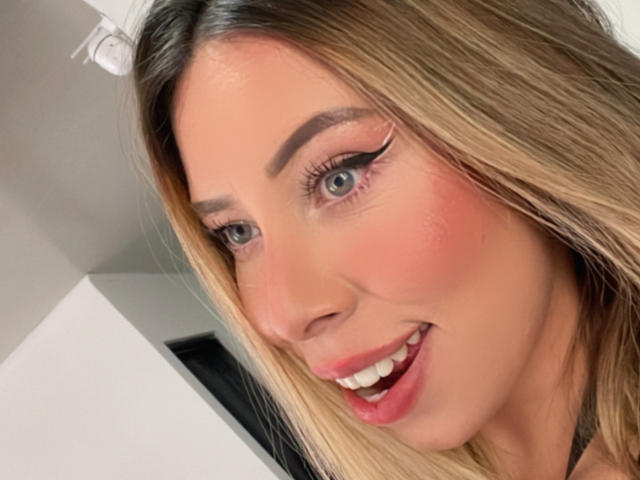 JennyLawry's face with blonde hair, blue eyes, pink blush, winged eyeliner, and a smile showing her pink lips and white teeth. She has a cute, attractive look.