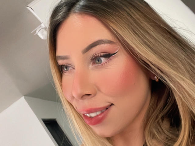 JennyLawry has blonde hair, blue eyes, and is smiling with pink lipstick. She has winged eyeliner and a slight blush on her cheeks. Her tits are not visible.