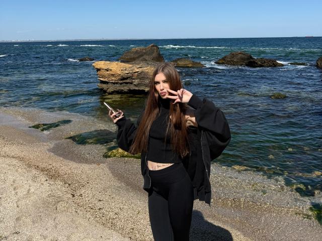 SensualMila mit langen, braunen Haaren, einem schwarzen Crop-Top und schwarzen Hosen, hält ein Telefon, macht ein Friedenszeichen und steht auf einem felsigen Strand mit Meer und Felsen im Hintergrund..