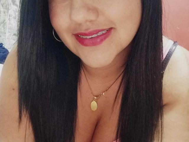 CarolinaConck with black hair, red lipstick, gold necklace, showing cleavage, small hoop earrings, smiling, slightly visible pierced teeth, wearing a pink and black striped top.
