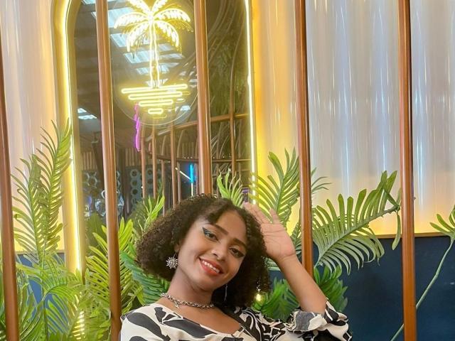 MeganWlliams with curly black hair, smiling, wearing a black and white patterned top, holding her hair, green plants, and neon lights in the background. She's got pretty earrings.