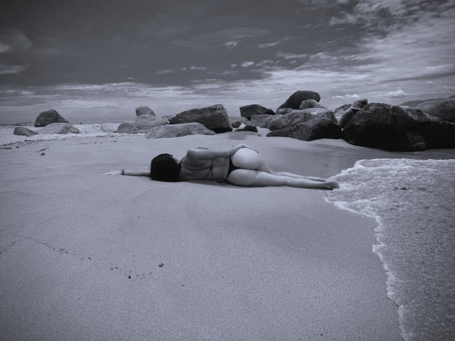 Jeanne Williams è sdraiata a faccia in giù su una spiaggia sabbiosa, indossando un bikini che a malapena copre il suo sedere e i suoi genitali, con le gambe distese e delle rocce sullo sfondo..
