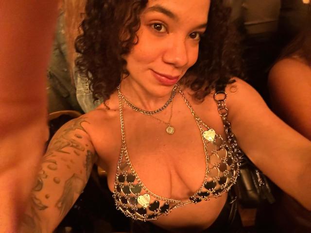 XFullOfLoadTransX with light brown skin, blonde hair in a ponytail, wearing a blue lace bra, smiling at the camera, showing small tattoos on her right arm.