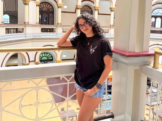 VivienneLaurent has curly black hair, wears a black t-shirt with a small pink bunny, blue denim shorts, and glasses, standing with one hand on her head.