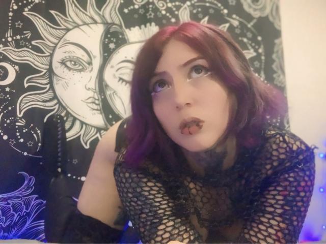Alice Calloway mit lila Haaren, schwarzem Netzoberteil und Zungenspalt, die mit großen Augen nach oben schaut, wobei Tattoos am Hals sichtbar sind. Im Hintergrund sind Gesichter von Sonne und Mond zu sehen..