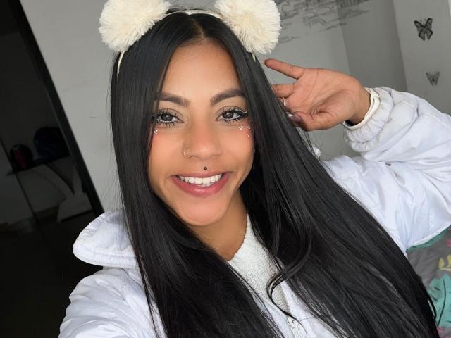 Amberv, con lunghi capelli neri, indossa orecchie da orso bianche e pelose, una giacca bianca, sorride, ha glitter negli occhi, un piercing al naso e una mano vicino all'orecchio, mostrando le sue unghie bianche..