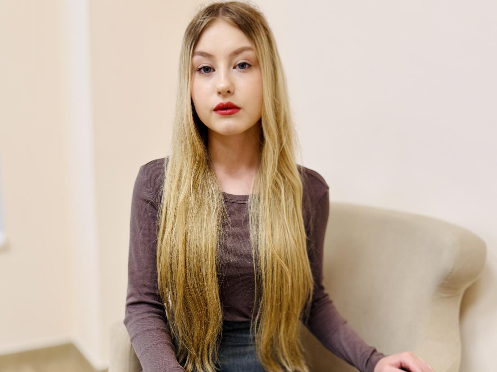 NevaBrowne, avec ses longs cheveux blonds, son rouge à lèvres rouge, portant un haut violet à manches longues, assise sur une chaise beige, a l'air légèrement ennuyée. Ses seins sont petits, son visage est joli..
