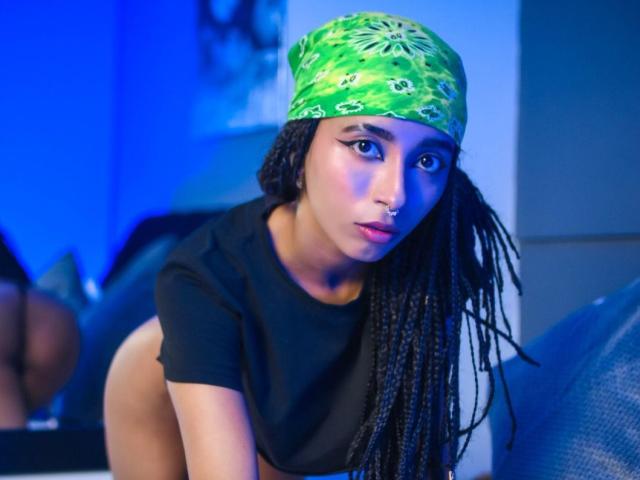 AgnieSoule, avec un bandana vert, un t-shirt noir et de longues tresses, regardant de manière séduisante l'appareil photo, son anneau de nez et son eye-liner ailé ressortant, avec un éclairage bleu en arrière-plan..