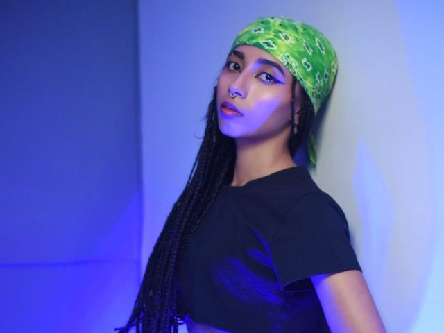 AgnieSoule, con trenzas largas, una bandana verde, una camisa negra, una luz azul y lápiz labial morado, apoyada contra una pared, mirando a la cámara con una expresión segura..