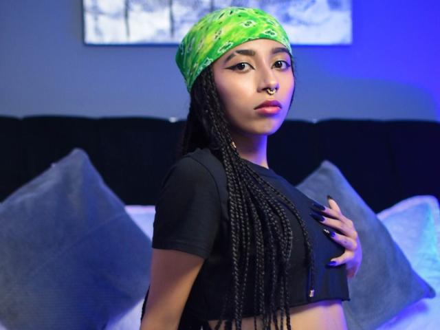 AgnieSoule, avec un bandana vert, un crop top noir et de longues tresses, touchant ses seins, avec un anneau de nez et des ongles bleus, se tient devant un canapé sombre..